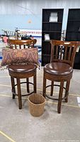 Two bar stools side by side with decorative pillow on left seat and wicker basket in front of stools