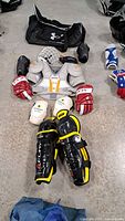 Photo showing arranged youth hockey protective gear including gloves, shin guards, elbow pads, shoulder pads, and helmet alongside a black Under Armour sports bag.