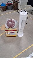 Image showing white tower air purifier fan with cord beside new Sunbeam vaporizer in box on concrete floor.
