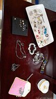 Photo showing various pierced earrings arranged on black card and white ceramic floral tray, along with bead bracelets and decorative ring holder dish on dark wooden surface.