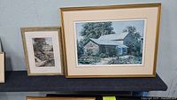 Two framed Keirstead prints displayed on a shelf against a gray wall, showing a larger landscape print with stone house and smaller house print beside it.