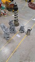 Five lawn ornaments displayed on concrete floor including lighthouse solar light and three child figurines with puppies plus metal watering can.