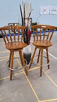 Two maple wood bar stools positioned side by side with a large vase holding dried flowers behind them.