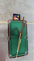 Overhead view of the miniature pool table showing green felt surface, two cue sticks, ball rack, and blue chalk blocks