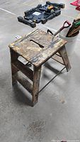 Wooden step stool with visible wear and paint splatters, standing on concrete floor with tools in background.