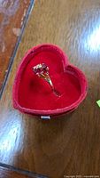 Gold plated ring with red ruby colored stones and clear accent stones in cluster design shown inside heart shaped red velvet box.