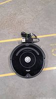 Top view of black iRobot Roomba vacuum with charging base and power cord on concrete floor.