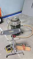 Photo showing Ridgid shop vacuum, work table, mitre box with saw, and trouble light lying on concrete floor against wall.