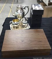 Silver plated teapot, cream and sugar set with a small tray, metal napkin holders stacked in boxes, and a closed wooden box on a black surface.