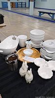Photo of white ceramic dishes including stacked bowls, a large curved serving bowl, white mugs, small serving bowls, and ceramic bird-shaped decorations arranged on a table.