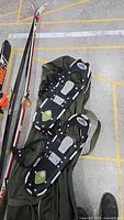 Photo showing a pair of black snowshoes with metal cleats and straps placed on a green bag along with one pair of cross-country skis.