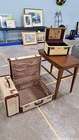 Photo shows two vintage luggage pieces - one open suitcase with brown ruched fabric lining and one smaller trunk-style suitcase with brown floral fabric lining - positioned next to a small wooden side table with tapered legs in medium brown finish.