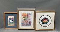 Three framed lithographs visible on a neutral gray background. Left print features a cow with heart graphic in a wooden frame, center shows flowers and jars with signature visible, right shows flower arrangement within a round mat and gold frame.