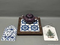 Grouped view of a purple teapot on a tiled wooden trivet, a blue and white ceramic tile, and a Christmas tree ceramic decoration.