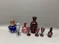 Full lot showing all colored glass items including blue bottle, gray vase, pink pitcher, ruby red decanter, four stem glasses, and small covered bottle