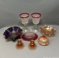 Collection of colored glass items including two clear stemmed glasses with red rims, purple iridescent bowl, beige dish, two pinkish-orange candle holders, and two covered dishes