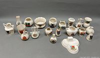 Full view of all 18 English porcelain souvenir trinkets arranged in rows on a flat surface showing various crests and shapes including jugs, bowls, mini vases, and pitchers.