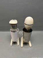 Photo showing two vintage electric drink mixers side by side, one branded Iona and the other Hamilton Beach, with their metal mixing cups attached.