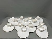 Arrangement of 13 white fluted plates paired mostly with cups inside each platter, showing the full set.