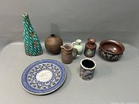 Seven pottery pieces placed on a flat surface including vases, pitchers, plate, and bowl in various colors and styles.
