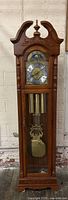 Front view of Ridgeway grandfather clock showing carved top, clock face, pendulum, and weights inside glass door.