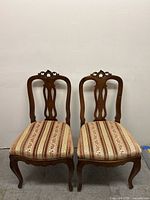 Pair of wooden dining chairs with upholstered patterned seats, carved backs, and curved legs, shown against white wall.