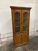 Full view of vintage colony maple corner cabinet showing two glass-paneled doors in upper section and two wooden doors in lower section with carved trim detail.