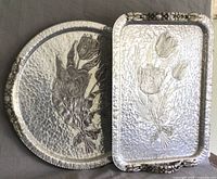 Two hand-wrought aluminium trays on grey background. Both show hammered surfaces with embossed tulip bunches and floral handles.