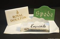 Front view of three vintage advertising store signs: Royal Doulton white ceramic, green ceramic Spode, and clear acrylic Exquisite sign.