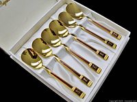 Six gold tone enamelled spoons with dark red handle detail arranged in white satin lined box.