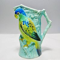 Front and side views of the colorful parrot jug showing detailed hand-painted design and aqua background with textured leaves and fruit.