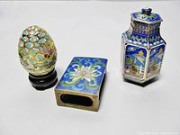 Front angled view of matchbox cover, egg-shaped figurine on stand, and hexagonal covered jar showing overall colors and details.