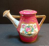 Front side view of the watering can shaped figural vase showing the hand-painted floral basket design on a pink maroon background with gold leaf accents on the body and spout.