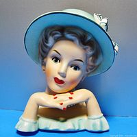 Front view of the head vase showing the woman's face, makeup, and crossed hands with red painted nails under a wide brimmed hat with checkered ribbon and bow.