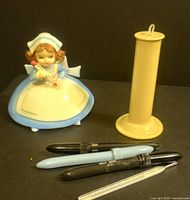 Photo of nurse figurine in blue dress holding thermometer, beige celluloid thermometer holder, and three vintage medical thermometers.