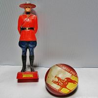 Front view of Mountie figurine standing upright next to round RCMP paperweight showing front illustrations and markings.