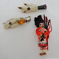 Photo showing all three items together: two Scottish grouse foot pins with fur and stone accents, and a miniature doll brooch dressed in traditional Scottish attire playing bagpipes.