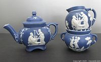 Front and side view of the blue jasper ware teapot, cream pitcher, and open sugar bowl with classical white relief decoration, set stacked for display.