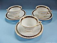 Front angled shot of three matching white porcelain cups and saucers with cobalt blue and gold decorative borders. Cups have flared shape and delicate gold accented handles.