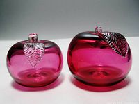 Front view of two cranberry glass apple paperweights showing their rounded shapes, red glass color, and clear glass leaves with veins.