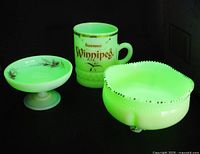Three uranium custard glass pieces displayed under black light showing their bright green fluorescence: Winnipeg souvenir mug, small pedestal dish with hand-painted roses, and a larger footed beaded bowl.