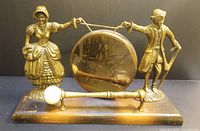 Front view of the vintage brass dinner gong held by a colonial couple on wooden base with mallet in front.