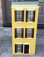 Front view of the yellow Victorian dollhouse showing six windows with black shutters, a black door, and a decorative white balustrade on the roof.