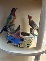 Three ceramic bird figures arranged on a white shelf: a bird-shaped planter with yellow flowers and two standing bird figures with pastel-colored gradient glazes.