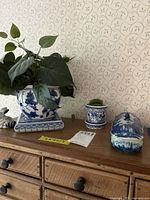 Four blue and white china pieces displayed on wooden dresser: large vase on square base with plant, small planter with moss, covered dish, and rabbit figurine.