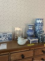 Wide shot showing all seven pieces arranged on a wooden surface against patterned wallpaper, includes vases, containers, and a porcelain bird figurine.