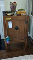 Front view of the wooden cabinet showing single door with decorative vent and latch, wood grain, and small trinket hanging on handle.