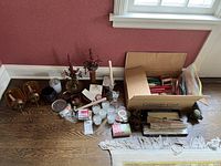 Wide view of metal and glass candle holders, taper candles, Yankee Candle tealights, and cardboard box with supplies all laid on wooden floor against red wall beneath window.