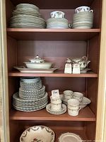 Full view of shelves showing stacked plates, bowls, teacups, saucers, serving dishes, and salt and pepper shakers arranged neatly.