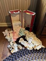 Red vintage doll trunk open, showing interior fabric lining, various vintage baby clothes, shoes, and Barbie doll outfits spread out on wooden floor.